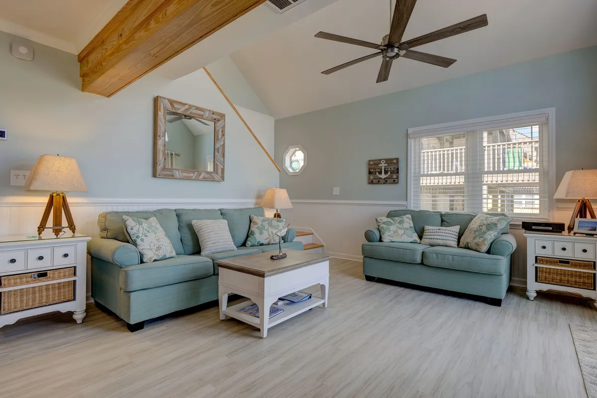 A spacious living area with light blue sofas, a white coffee table, wood-beamed ceiling, and soft natural light. The inviting layout is perfect for relaxing, reading, or gathering with guests.