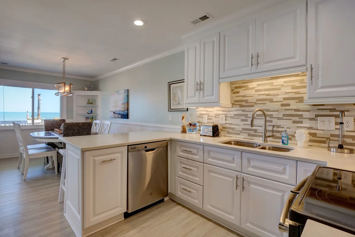A bright, cheerful kitchen and dining area with a kitchen island, modern stainless steel appliances, decorative wall art, and a dining table positioned beside large windows offering a direct ocean view. The space balances functionality with a sense of togetherness.