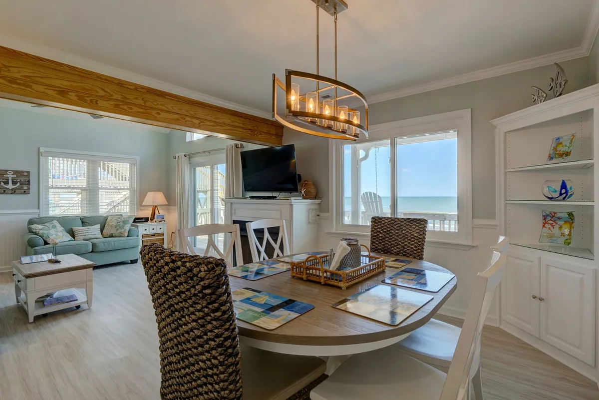 A cozy, open-concept interior space featuring a dining table with decorative placemats, a comfortable teal couch, large windows showcasing ocean views, and a chandelier above the table. The scene captures the ideal spot for families to regroup after exploring, sharing meals, and recounting the day's adventures in a welcoming, light-filled setting.