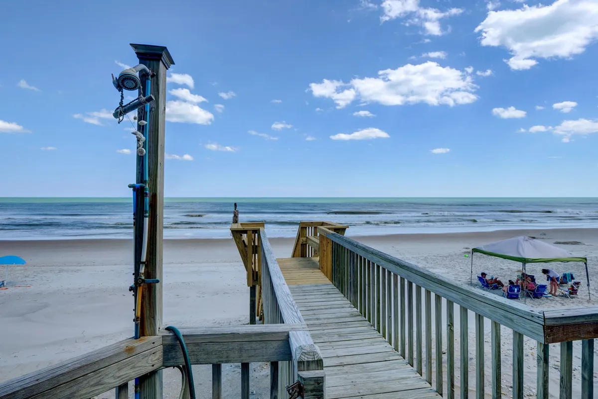 A wooden boardwalk stretches from the property toward the open beach, with the ocean glistening in the background. People relax under a beach umbrella nearby, while others stroll along the sand, swim, or enjoy a picnic.