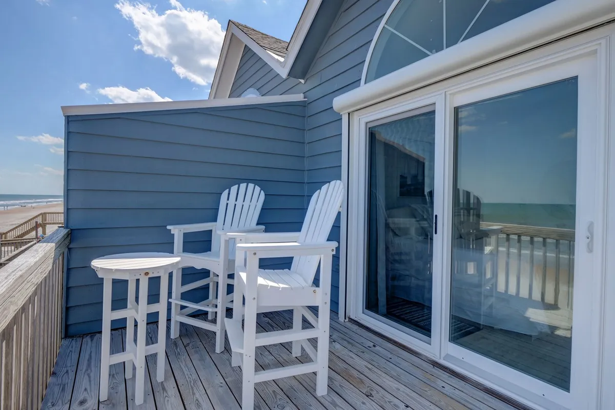 A close-up of two white Adirondack chairs and a small table on a deck, with a sliding glass door behind and a sweeping beach view ahead. The setup hints at intimate morning coffees or quiet sunset moments.