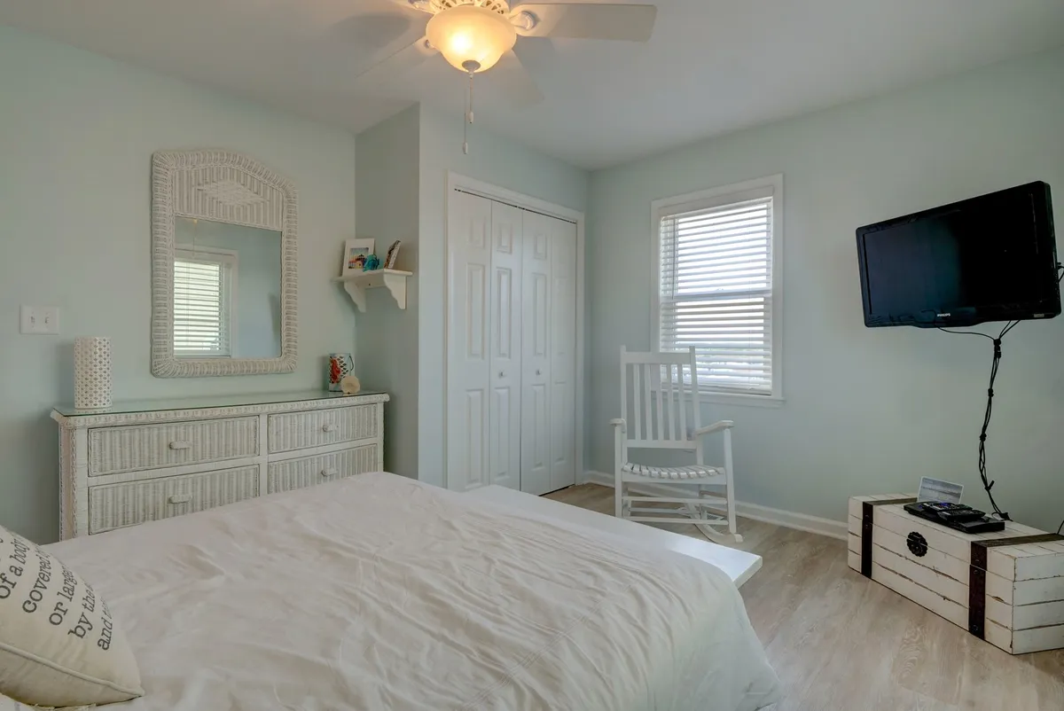 A cozy bedroom with a bed dressed in white linens, a classic rocking chair, a dresser with a mirror, and a wall-mounted TV. The soft lighting and simple decor create a restful and homey retreat.