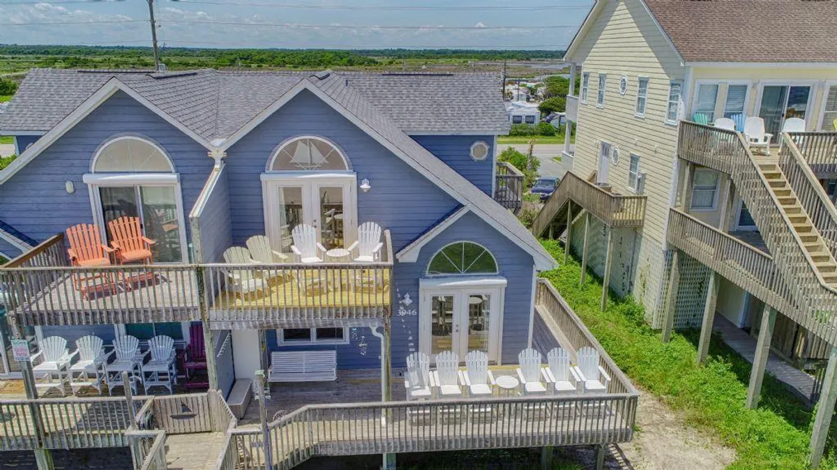 A bright exterior view of the blue house, highlighting a deck with white chairs and glimpses of adjoining homes. The atmosphere is cheerful and relaxed, suggesting outdoor amenities such as an outdoor shower might be nearby, while showcasing the private home setting.