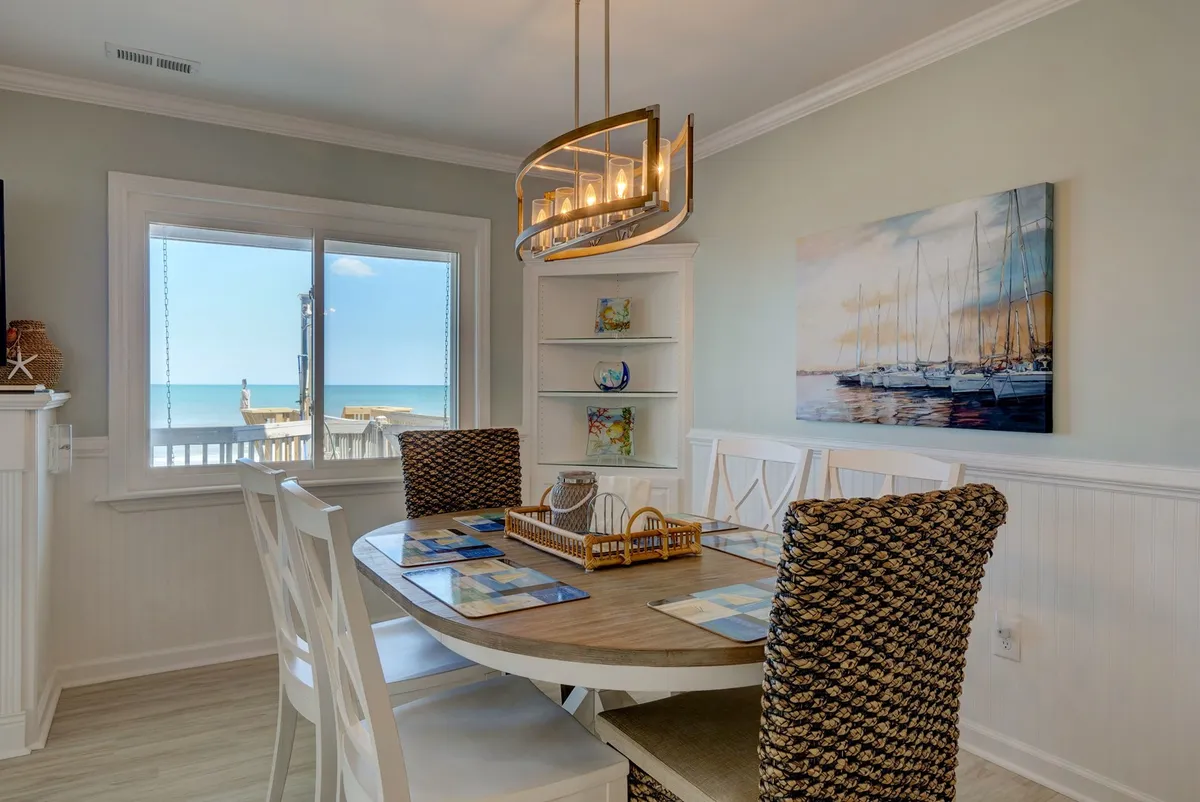 A cozy dining space with a table set by a large window overlooking the ocean, adorned with coastal artwork and decorative shelves. The atmosphere feels relaxed and coastal, perfect for enjoying coffee at sunrise or a sunset toast.
