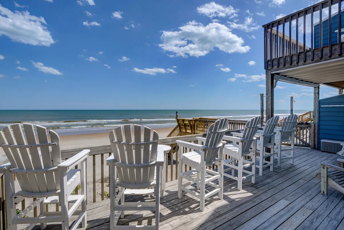 The hero image showcases a spacious beachfront deck overlooking the shimmering Atlantic Ocean, with inviting beach chairs positioned to soak in the panoramic views. The scene features clear blue skies, direct access to soft sands, and a tranquil atmosphere perfect for relaxing, social gatherings, or watching stunning sunrises and sunsets.