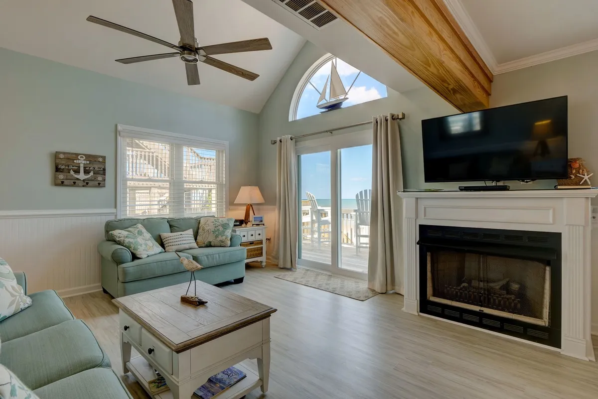 A cozy living room with a plush sofa facing a modern fireplace and a 50-inch TV, flanked by large windows that let in abundant natural light and offer glimpses of the ocean beyond. The room is accented by a ceiling fan and relaxed décor.