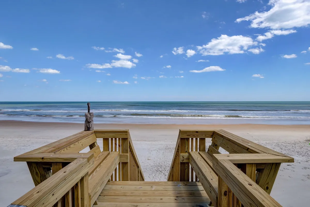 A tranquil beachside scene featuring wooden stairs leading down to the sand, with an expansive ocean view under a cloud-dappled sky. The image evokes the allure of the coast and the promise of beach activities like walking, sunbathing, shell collecting, and sandcastle building, aligning with the section's emphasis on family fun both on and off the sand.