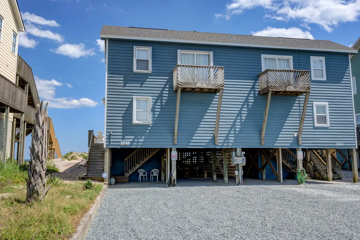 An exterior shot of the blue house with balconies and a gravel driveway. Two white chairs sit outside, providing a welcoming touch. The covered area beneath the balcony suggests a sheltered space, potentially used as a garage or for easy unloading.