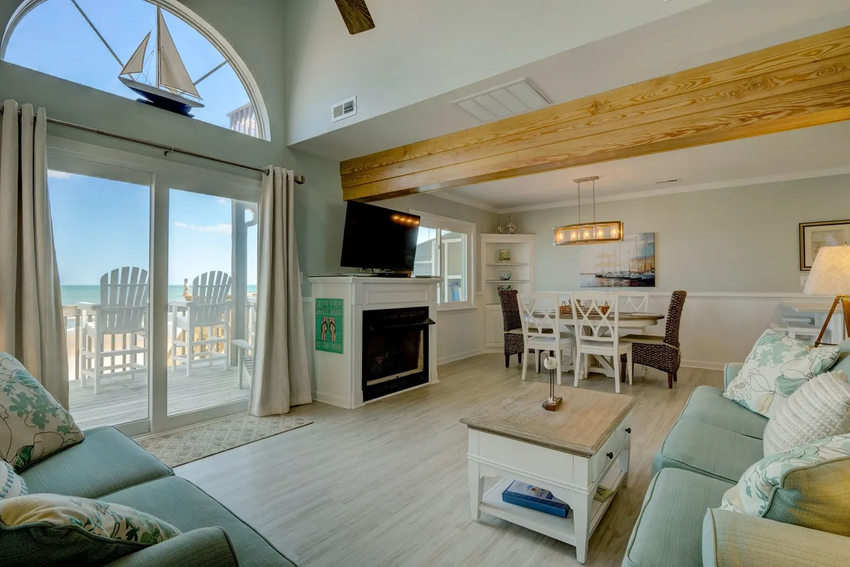 A welcoming open-concept living and dining area with large windows framing an expansive ocean view. The space features a comfortable seating area with plush sofas and a decorative sailboat, a dining table set for gatherings, and a cozy fireplace, creating a bright and communal atmosphere.