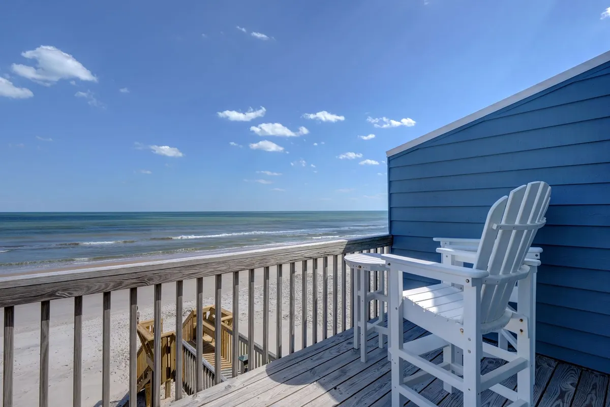 An exterior shot capturing the beach and ocean in the background, with deck chairs placed on a wooden deck area under a wide blue sky. The setup invites guests to unwind, sip drinks, or read while watching the sunrise or stargazing at night.