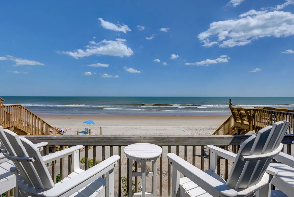 A serene wooden deck furnished with Adirondack chairs overlooks the ocean, with gentle waves visible just beyond the railing. The early or late sunlight casts a warm glow over the scene, inviting relaxation and reflection.