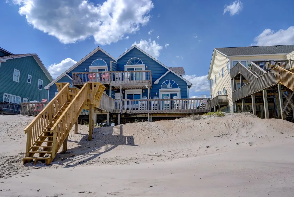 A cheerful scene featuring a blue house with prominent balconies and a wide wooden staircase leading directly to the deck. The sandy area surrounding the house hints at easy beach access and abundant outdoor social space, with the balconies ideal for viewing sunrises or sunsets.
