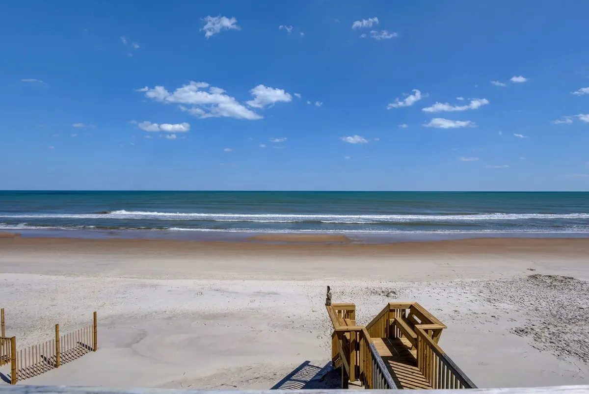 A tranquil view of the beach and ocean, with wooden stairs leading down to the sand. The scene is bathed in natural light, suggesting the perfect setting for morning walks or sunset relaxation. The image emphasizes openness and a direct connection to the outdoors.
